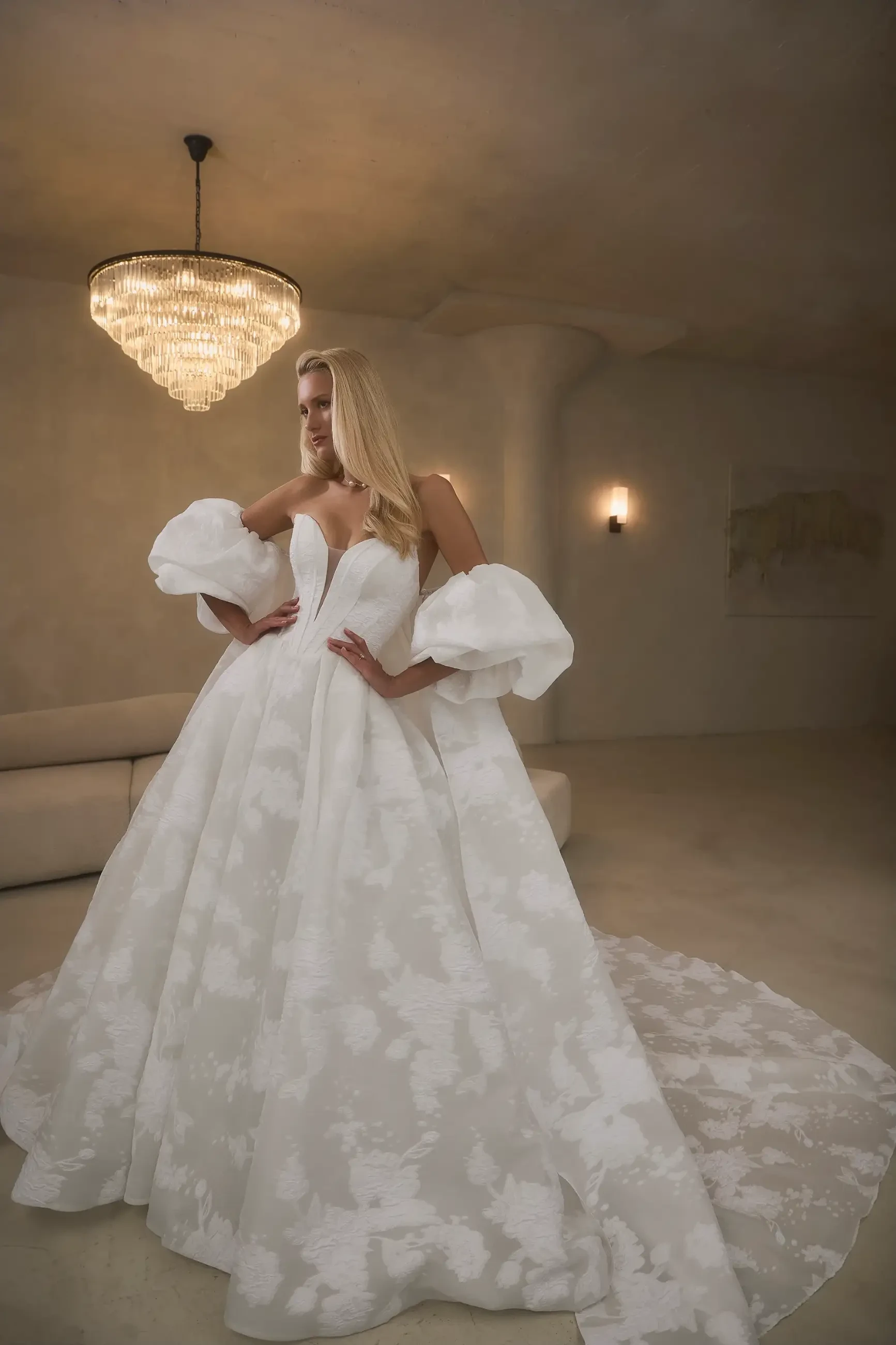 A woman wearing a white ball gown with puffed sleeves, standing elegantly in a softly lit room.