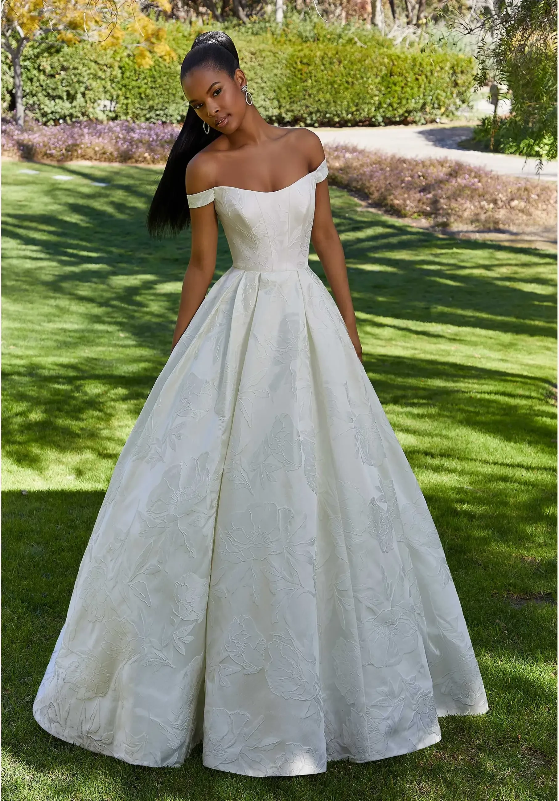 A woman stands in a garden wearing an elegant off-the-shoulder white ball gown, with her hair styled sleekly back.