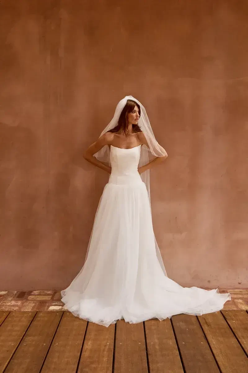 A woman in a white wedding dress and veil stands against a textured brown wall.