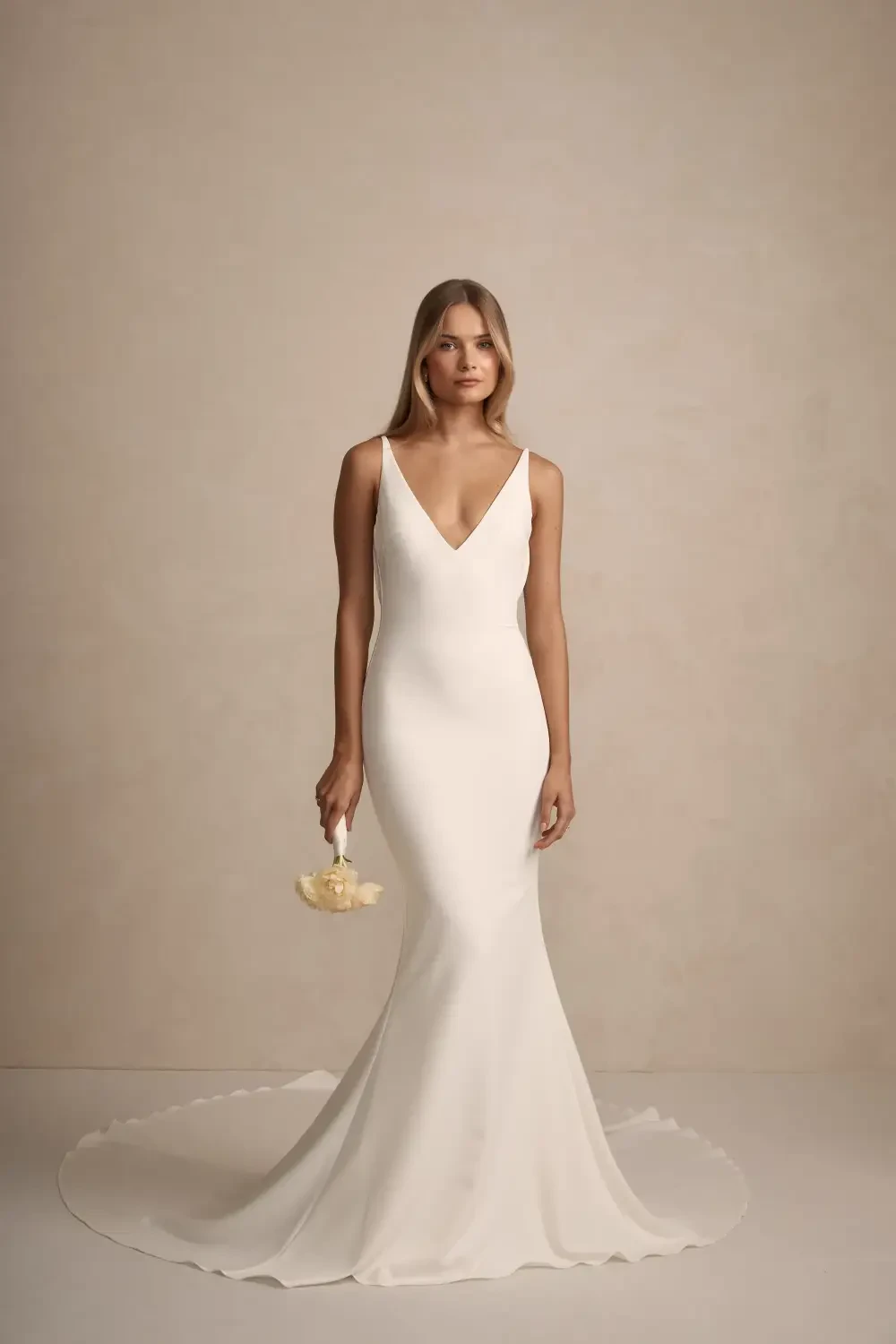 A model is wearing a sleek, white wedding dress with a deep V-neckline, holding a small bouquet of flowers, against a neutral background.