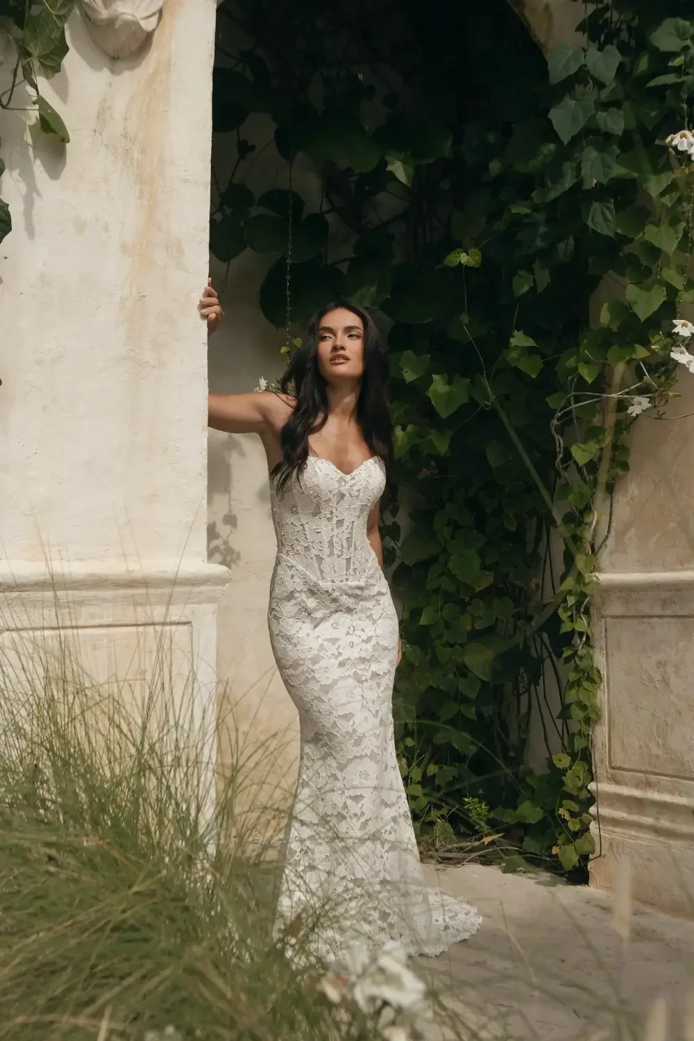 A woman in a fitted, lace wedding dress standing in a doorway surrounded by greenery.
