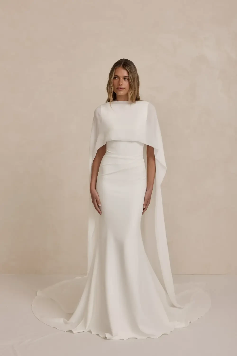 A woman wearing a elegant white gown with a cape against a neutral background.