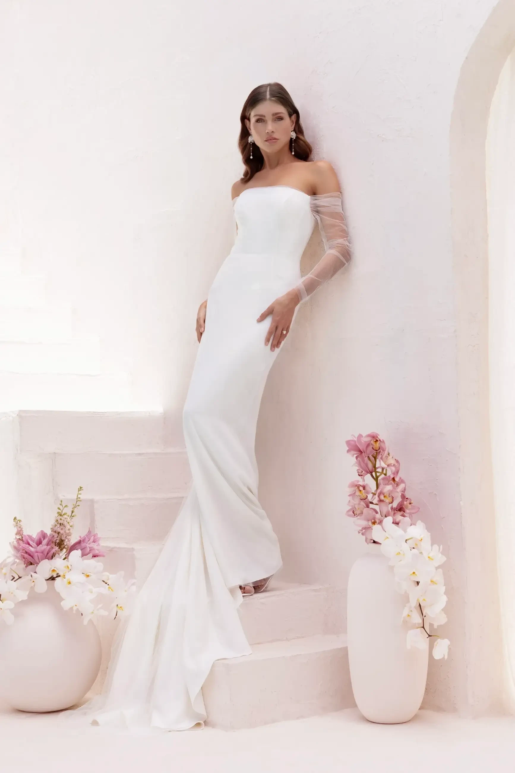 A model in a white off-the-shoulder wedding dress stands on a staircase with flowers in vases beside her.