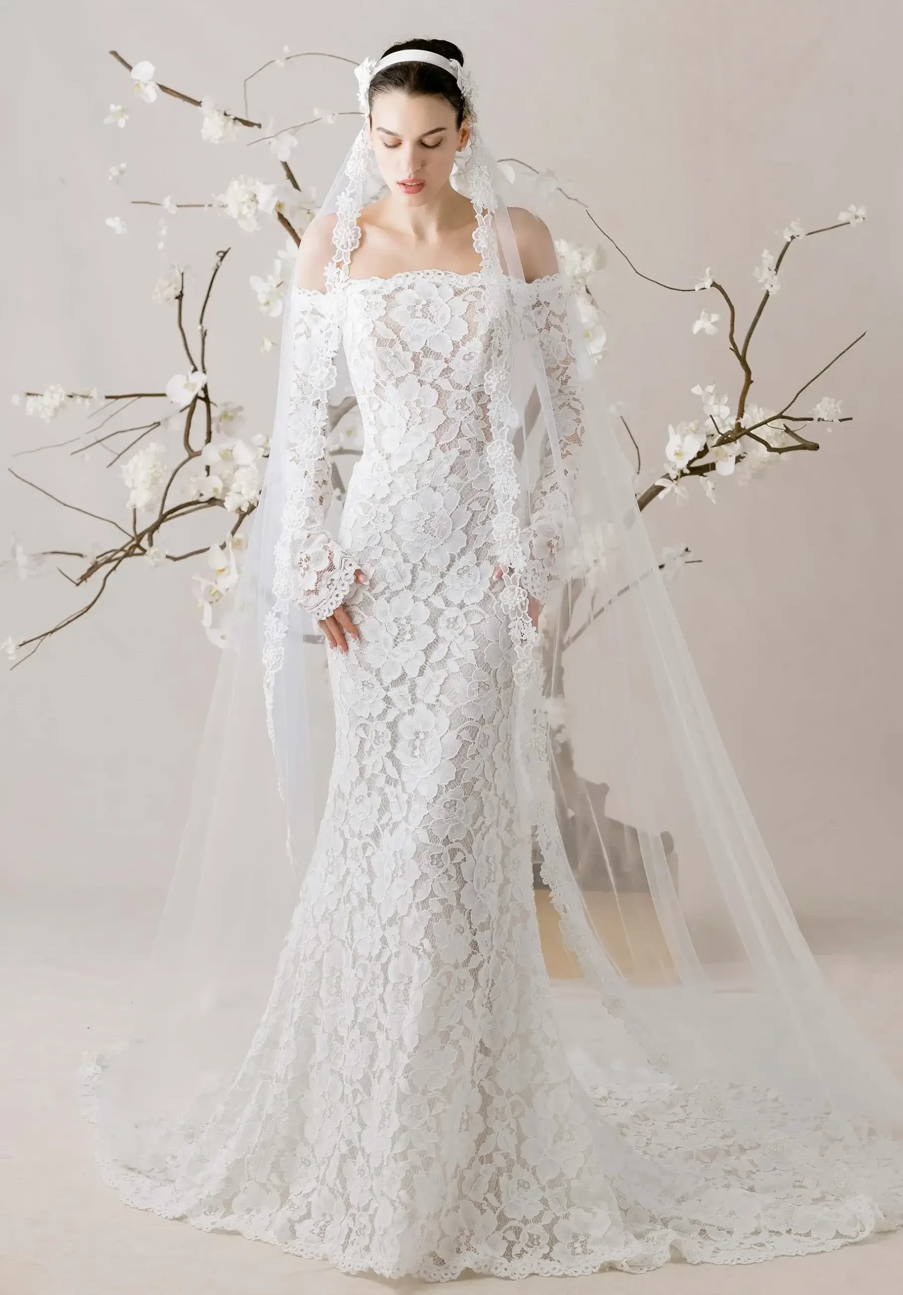 A model wearing an intricate white lace wedding dress with long sleeves and a flowing veil, standing in front of a minimalist backdrop adorned with branches and blossoms.