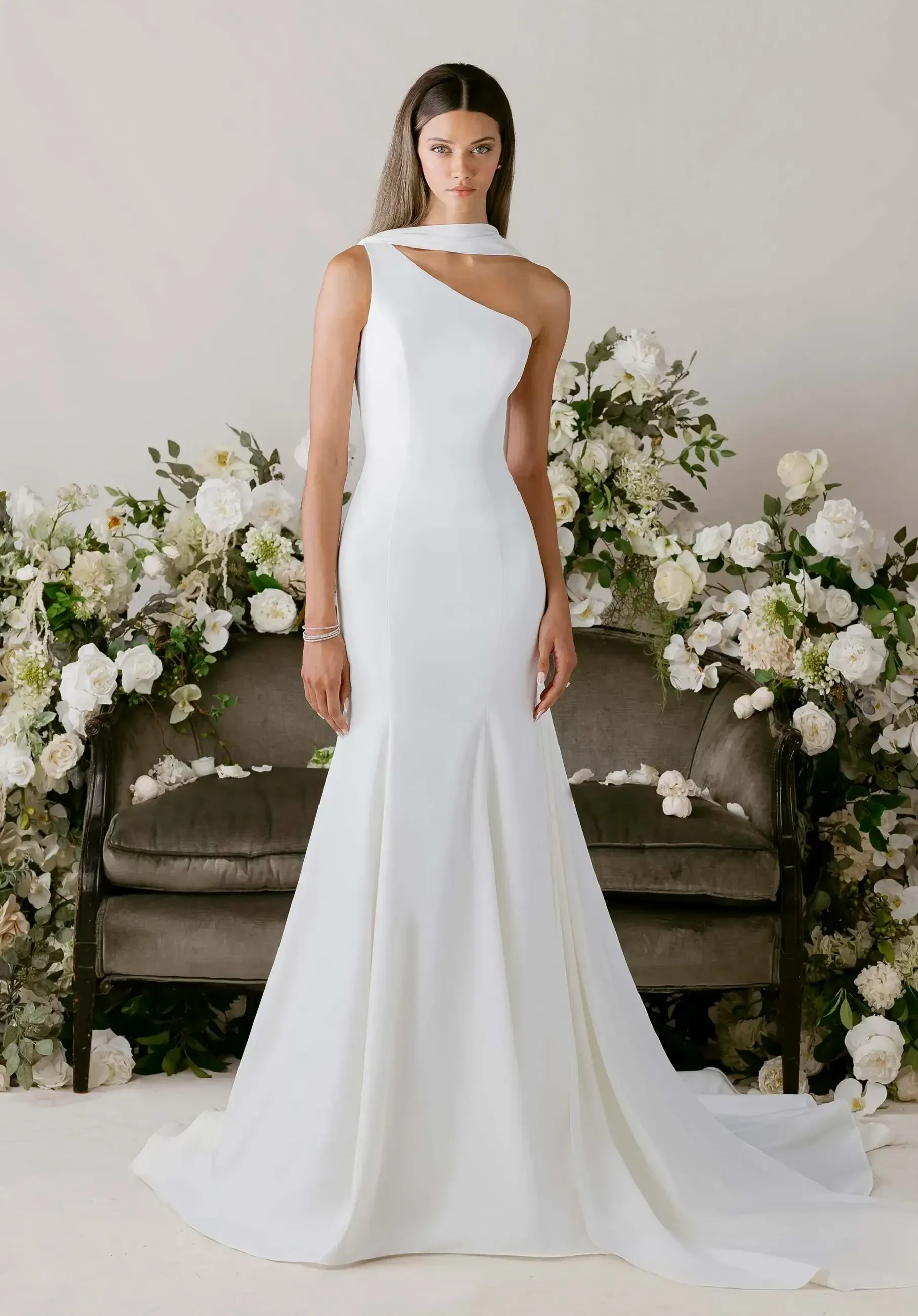A model wearing a fitted white gown with one shoulder design, standing in front of a floral arrangement and an elegant sofa.