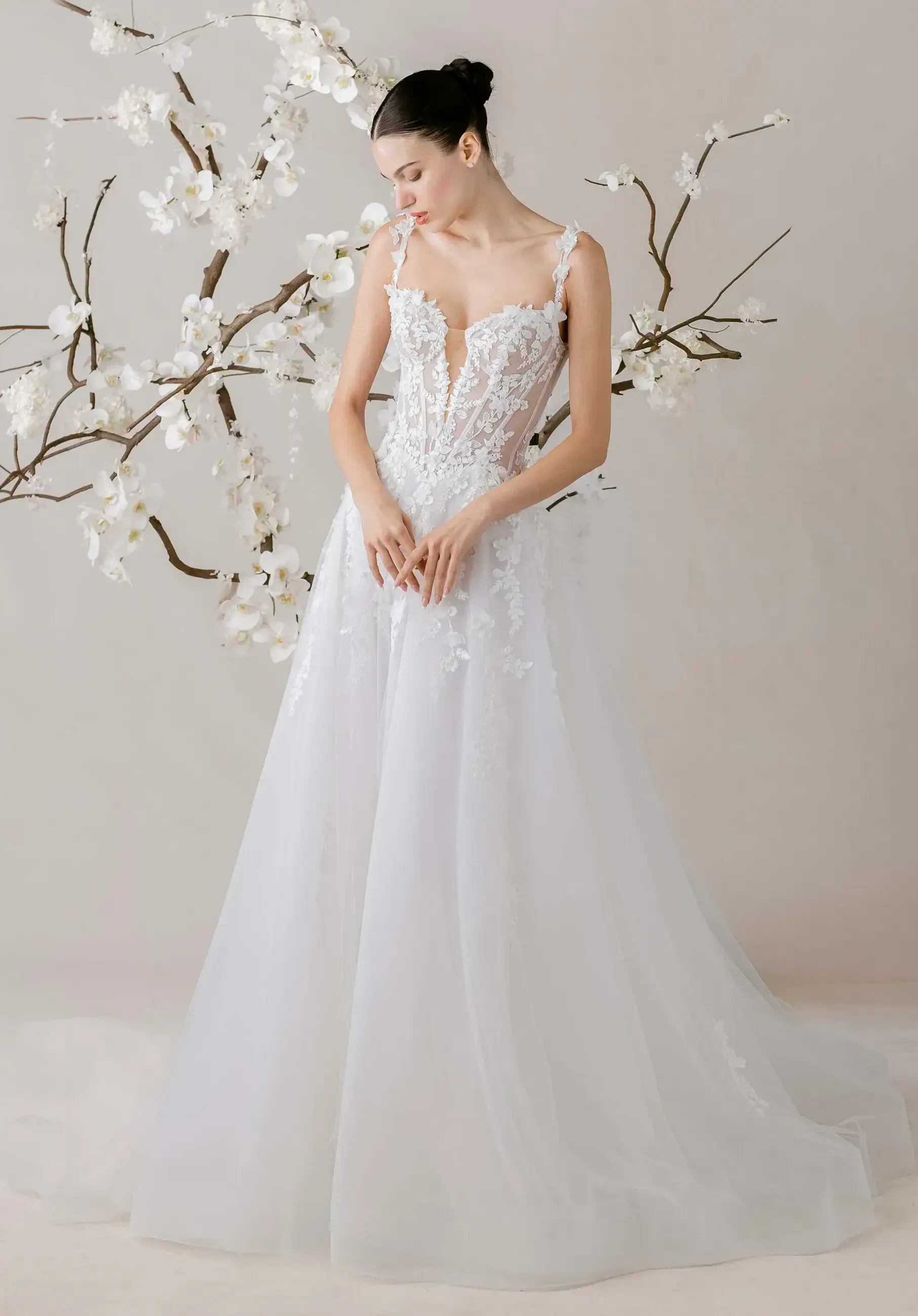 A model wearing a stunning white wedding gown, featuring intricate lace details and a flowing skirt, stands gracefully in front of a backdrop with blooming branches.